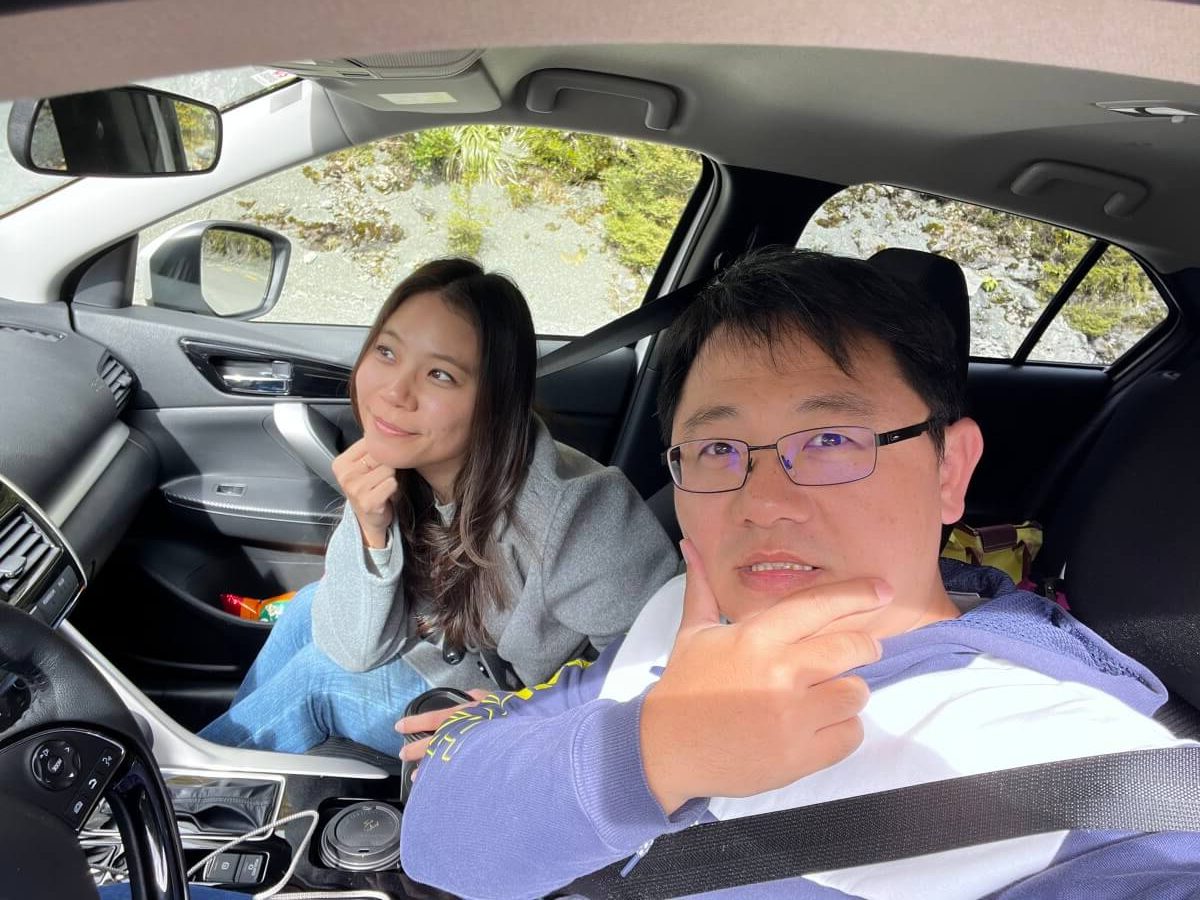 一男一女坐在車內，男子戴著眼鏡坐在駕駛座，女子坐在副駕駛座，兩人面向鏡頭，車窗外可見樹木和岩石。