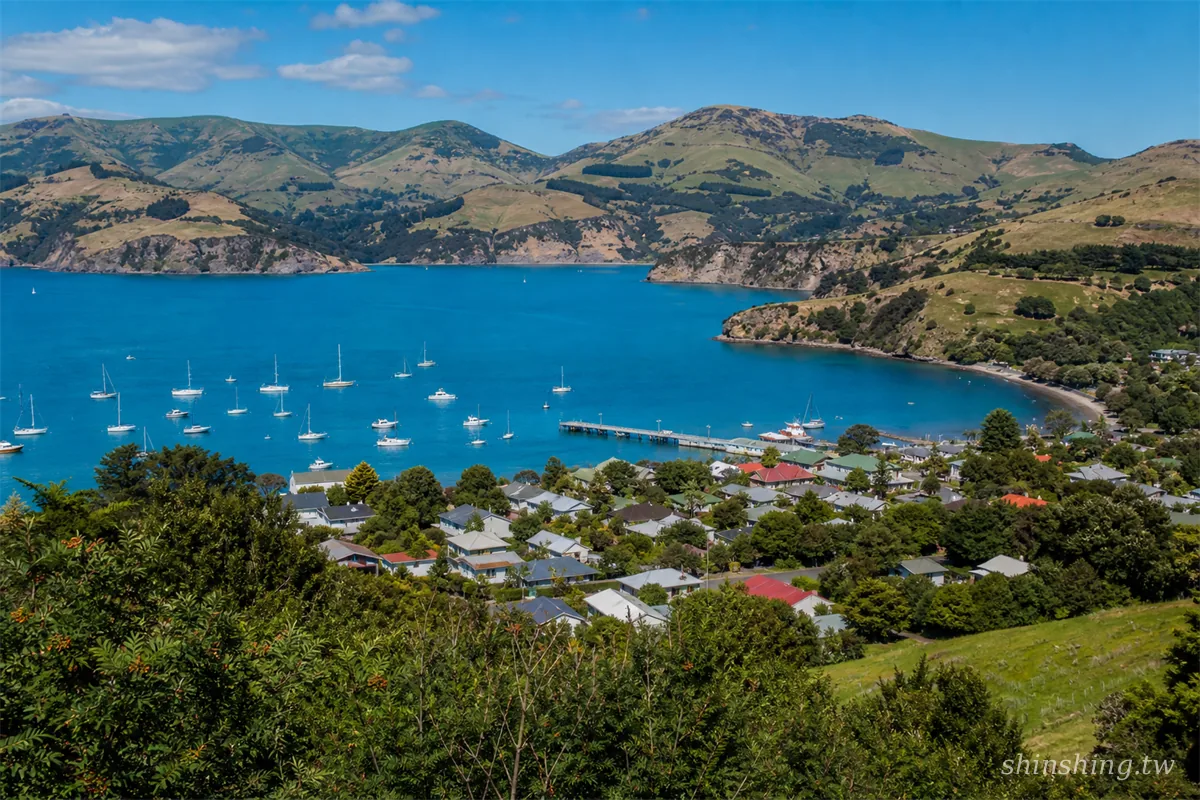 阿卡羅阿 Akaroa Harbour 法式小鎮全景，深藍港灣與火山峭壁環抱，白色帆船點綴水面