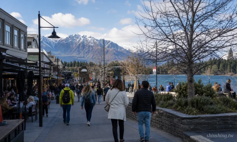 皇后鎮 Lake Wakatipu 湖畔步道一景，遠方 Remarkables 山脈雪頂倒映在湖面，湖邊綠樹與木製長椅