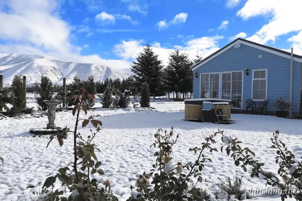 Kahurangi Cottage 的淺藍色小屋在雪後晴天下,屋外放著一座木製戶外熱水桶,遠方是覆雪的山脈