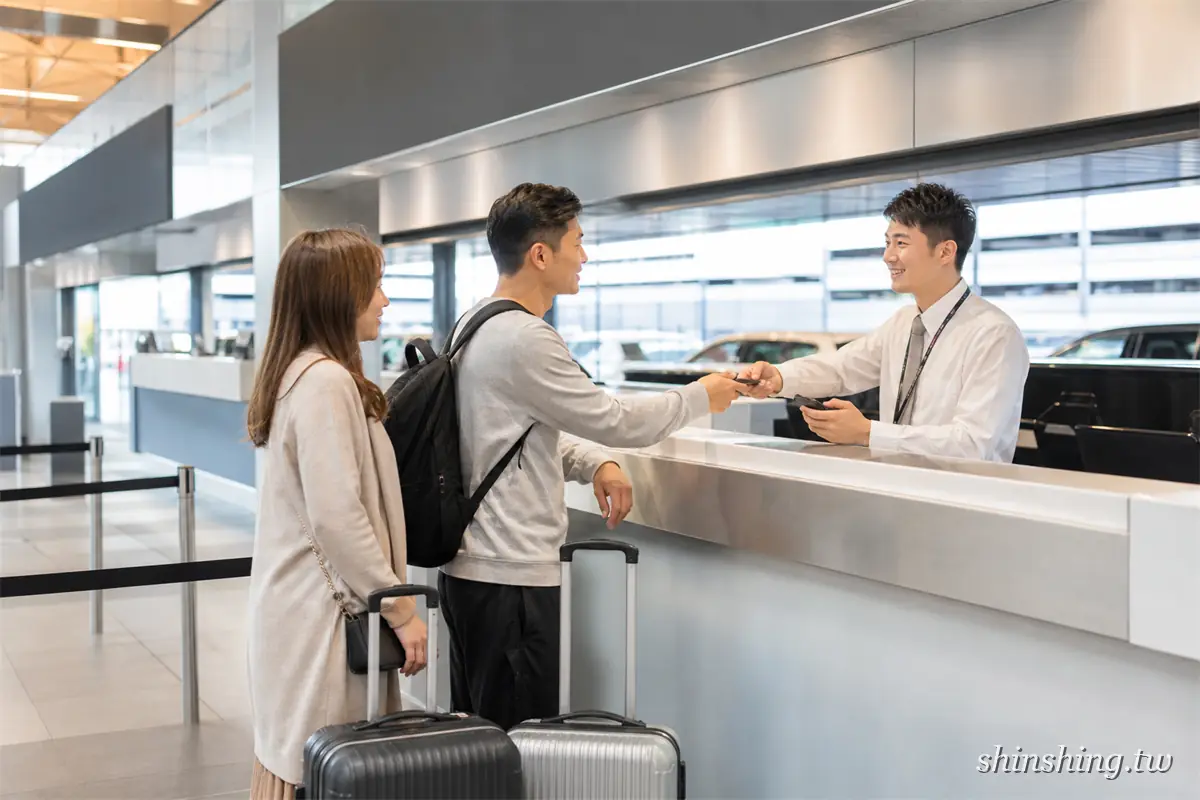 兩位旅客拖著行李箱在奧克蘭機場租車櫃檯辦理手續,櫃檯人員遞出車鑰匙,後方玻璃外就是待取的車輛