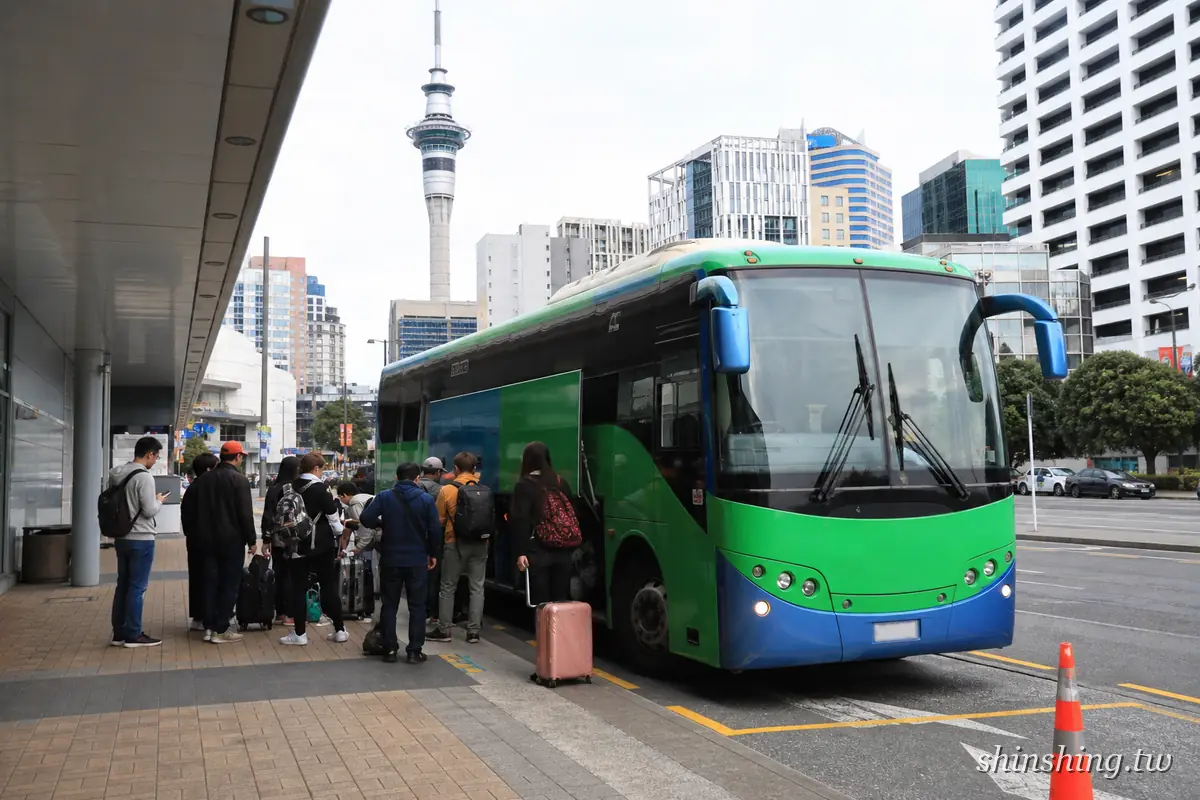 InterCity 巴士停靠奧克蘭 SkyCity 上車點,乘客拖行李排隊上車,背景是天空塔