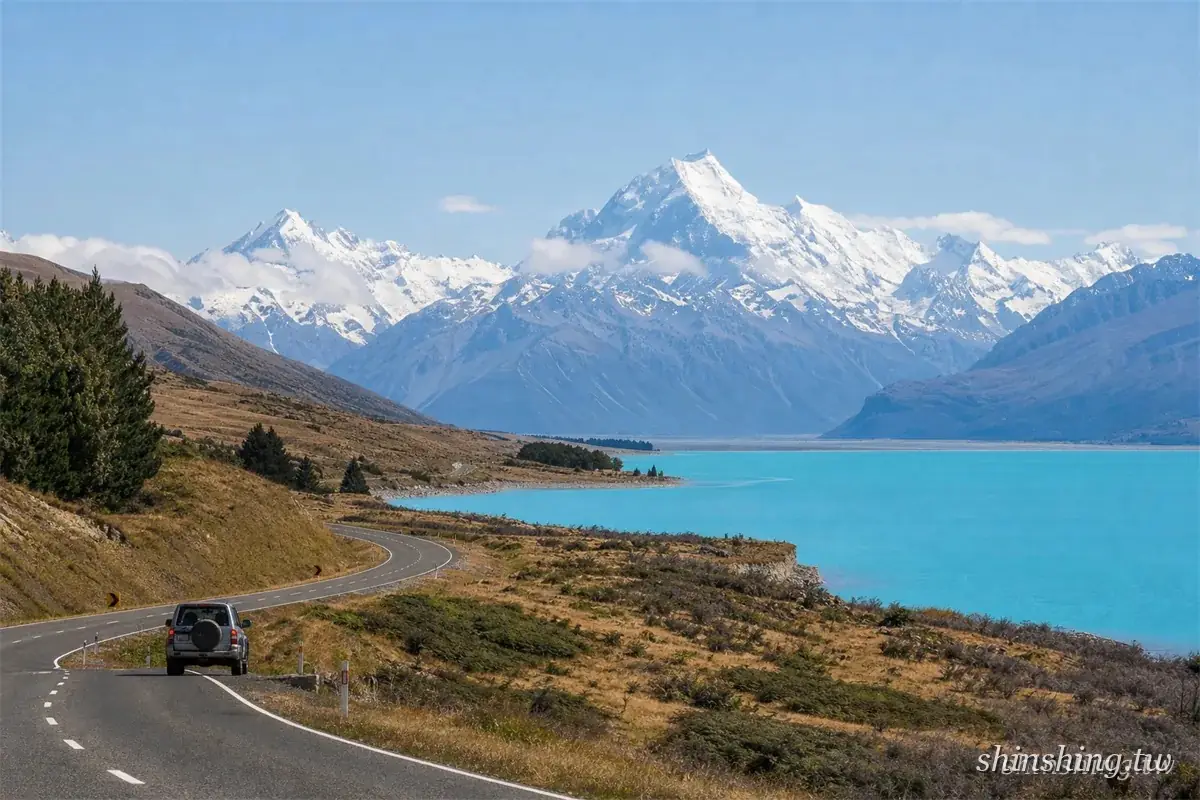 一輛休旅車行駛在 80 號公路上，右側是冰川藍的普卡基湖，正前方是覆雪的庫克山群峰
